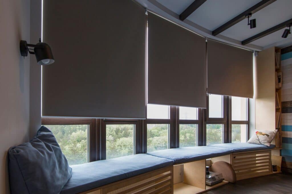 A cosy window seat with a blue cushion, featuring roller blinds and a view through the window.